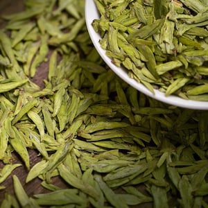 Puede incluir: Hojas de té verde sueltas en un tazón blanco y esparcidas sobre una superficie de madera.
