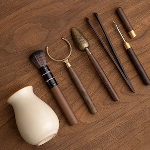Könnte beinhalten: Ein Set aus sechs Holz-Teewerkzeugen, darunter eine Bürste, eine Schaufel, eine Pinzette, ein kleiner Löffel und eine kleine Gabel. Die Werkzeuge sind alle aus dunklem Holz mit Messingakzenten.