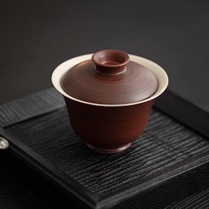 Könnte beinhalten: Eine braune Teetasse aus Keramik mit weißem Rand und Deckel. Die Teetasse steht auf einem schwarzen Holztablett.