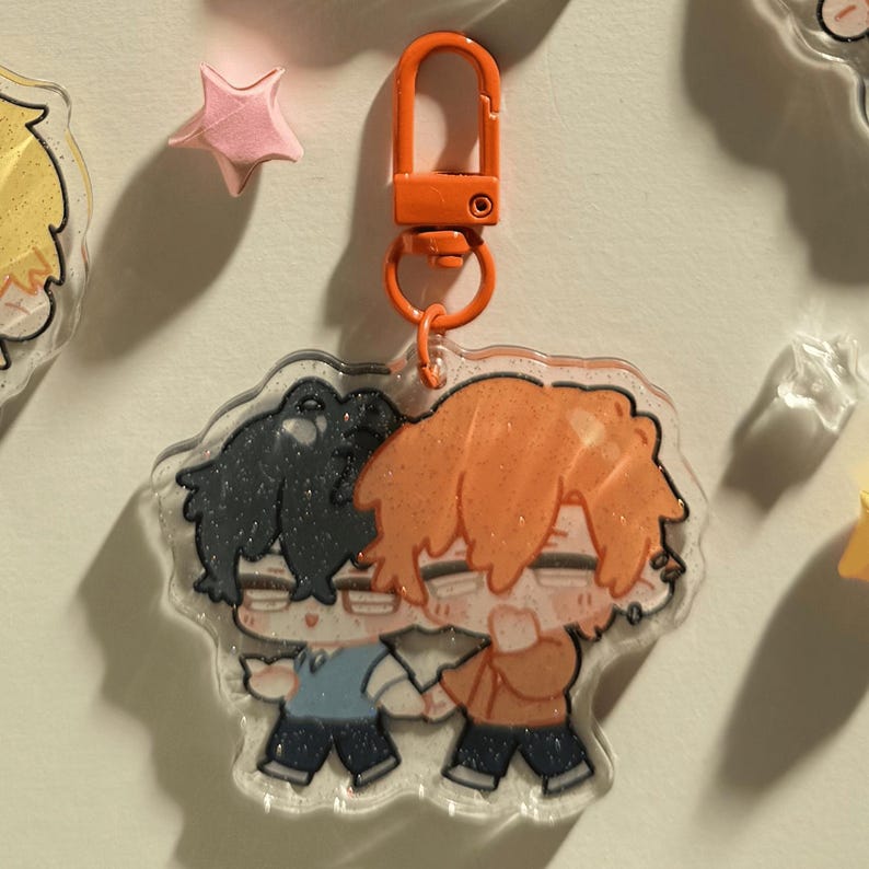Op de afbeelding: Een heldere acryl sleutelhanger met een cartoon illustratie van twee personages die elkaar omarmen. De sleutelhanger heeft een oranje sluiting en ring. De personages hebben zwart en oranje haar, gekleed in blauwe en oranje outfits. Een roze stervormige decoratie is zichtbaar.