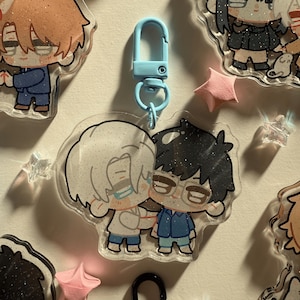 Op de afbeelding: Een heldere acryl sleutelhanger met twee cartoonfiguren die elkaar omarmen. De ene figuur heeft wit haar, de andere heeft zwart haar en een bril. De sleutelhanger heeft een lichtblauwe sluiting en is omgeven door andere bedels en stervormige decoraties.
