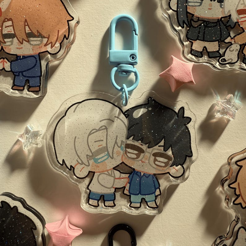 Op de afbeelding: Een heldere acryl sleutelhanger met twee cartoonfiguren die elkaar omarmen. De ene figuur heeft wit haar, de andere heeft zwart haar en een bril. De sleutelhanger heeft een lichtblauwe sluiting en is omgeven door andere bedels en stervormige decoraties.
