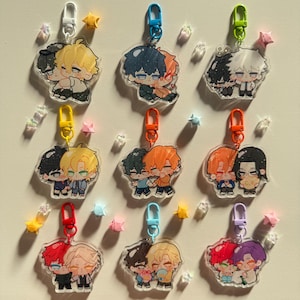 Op de afbeelding: Een verzameling anime-stijl acryl sleutelhangers met paren van knuffelende personages met verschillende haarkleuren. Elke sleutelhanger heeft een verschillend gekleurde sluiting, waaronder wit, blauw, groen, geel, oranje, rood en paars. Ster- en edelsteenachtige decoraties.