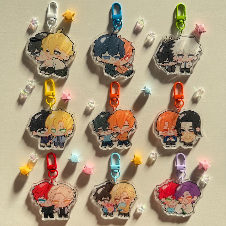 Op de afbeelding: Een verzameling anime-stijl acryl sleutelhangers met paren van knuffelende personages met verschillende haarkleuren. Elke sleutelhanger heeft een verschillend gekleurde sluiting, waaronder wit, blauw, groen, geel, oranje, rood en paars. Ster- en edelsteenachtige decoraties.