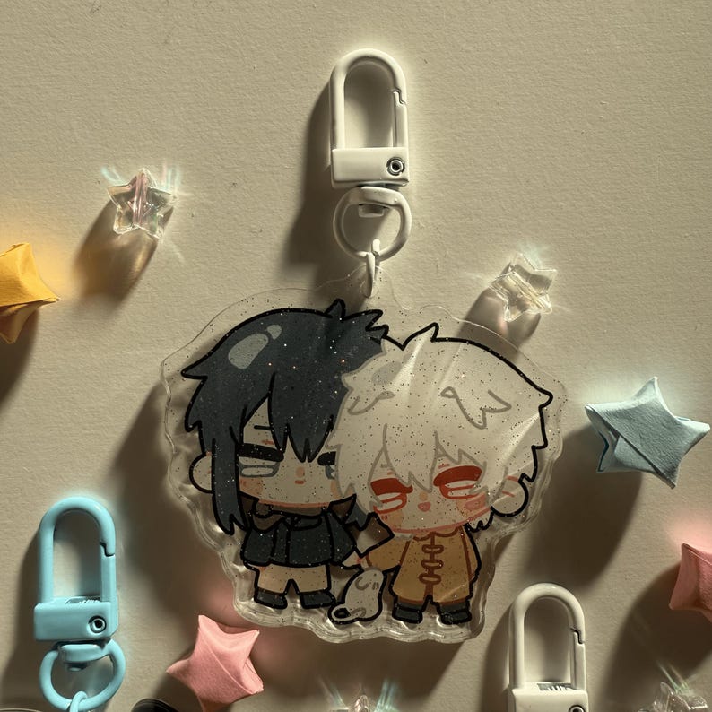 Op de afbeelding: Een heldere acryl bedel met twee anime-achtige personages die elkaars hand vasthouden. De ene heeft zwart haar en een zwarte jas, de andere heeft wit haar en een oranje outfit. De bedel is bevestigd aan een witte sluiting.
