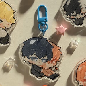 Op de afbeelding: Een heldere acryl sleutelhanger met een blauwe sluiting, met twee anime-achtige personages die elkaar omarmen. De sleutelhanger is versierd met glitter. Andere vergelijkbare sleutelhangers en stervormige decoraties zijn ook zichtbaar.