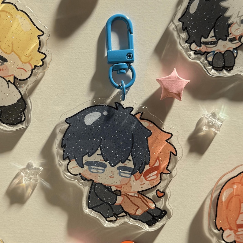 Op de afbeelding: Een heldere acryl sleutelhanger met een blauwe sluiting, met twee anime-achtige personages die elkaar omarmen. De sleutelhanger is versierd met glitter. Andere vergelijkbare sleutelhangers en stervormige decoraties zijn ook zichtbaar.