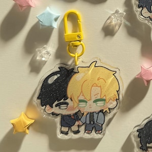 Op de afbeelding: Een heldere acryl sleutelhanger met een cartoon illustratie van twee figuren die elkaar omarmen. De figuren hebben zwart en blond haar, dragen pakken en houden elkaars hand vast. De sleutelhanger heeft een gele sluiting en is omgeven door origami sterren.