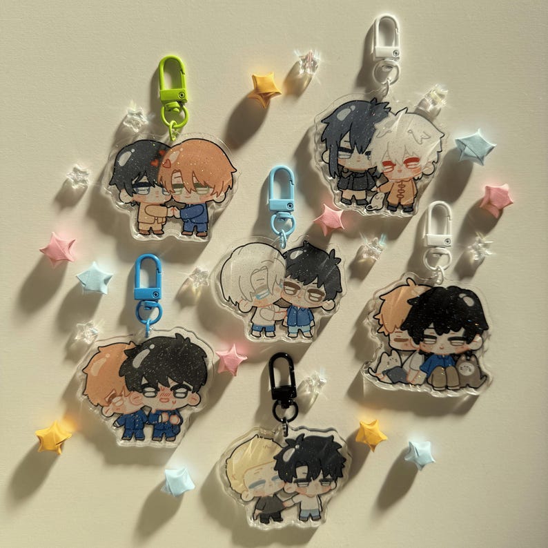 Op de afbeelding: Een verzameling heldere acryl sleutelhangers met cartoon illustraties van knuffelende koppels. Elke sleutelhanger heeft een verschillend gekleurde sluiting en is omgeven door stervormige en kristalachtige decoraties.