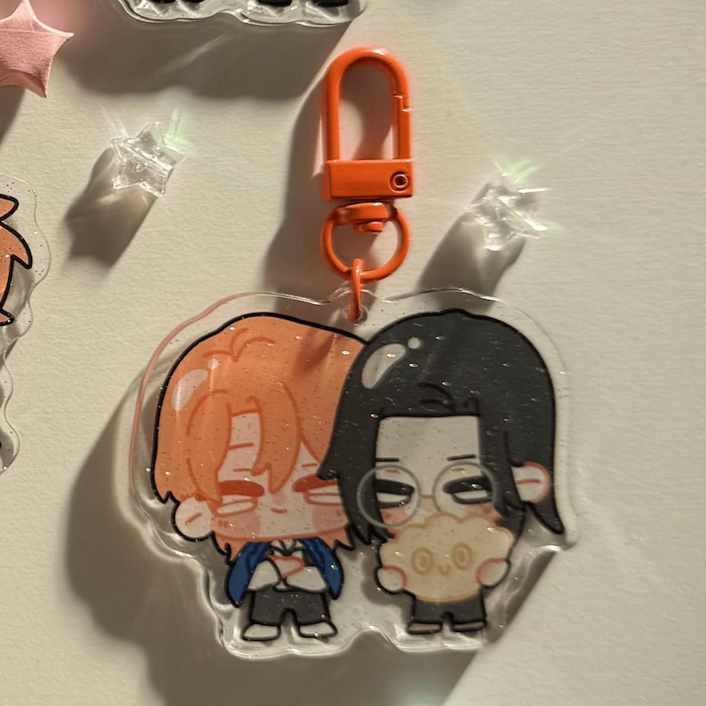 Op de afbeelding: Transparante acryl bedel met twee anime-stijl personages, een met oranje haar, de ander met zwart haar. De bedel heeft een oranje sluiting en is versierd met glitter. De personages dragen blauwe en zwarte outfits.