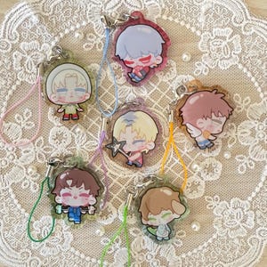 May include: A collection of seven acrylic charms featuring cartoon characters with various hair colors and outfits. Each charm has a silver-colored clasp and a colorful lanyard. The charms are displayed on a white lace surface.