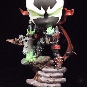 May include: A stylized Spawn figurine featuring a large head, a black mask with white eye markings, and a red cape. The character holds a sword with green glowing accents, stands on a stone-like base, and wears spiked armor with chains.