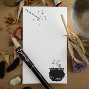 May include: A white lined notepad with a black pen and a black and white illustration of a magic wand and a cauldron with bubbles. The notepad has a black and white illustration of a magic wand and a cauldron with bubbles.