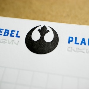 Op de afbeelding: Een wit notitieboek met een zwart Rebel Alliance-symbool en de woorden "Rebel Plans" in blauw.