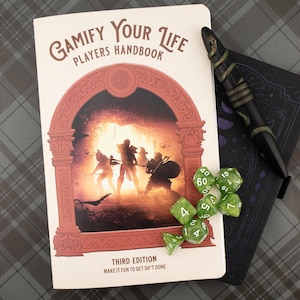 May include: A softcover book titled Gamify Your Life Players Handbook Third Edition Make It Fun To Get Sh*t Done, featuring a fantasy illustration of adventurers in an archway, accompanied by green polyhedral dice and a black pen.