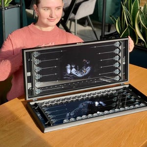 May include: Backgammon board with a black and silver design. The board is open, revealing a black image of a cat. The game board is set on a wooden table.