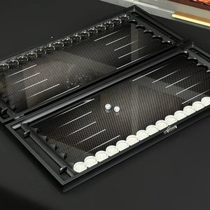 May include: A black backgammon set with a carbon fiber design. The set is open, revealing white and clear game pieces. The board is rectangular with a series of white and clear pieces. The set is displayed on a black surface.