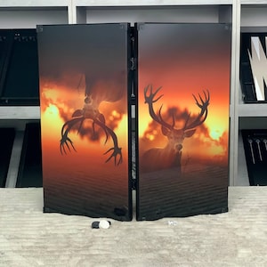 Peut inclure: Un plateau de backgammon pliant orné d'un graphisme contrasté représentant un cerf aux grands bois sur un fond de coucher de soleil orange et jaune vif. Le plateau est noir avec une finition brillante, montrant le cerf droit et inversé.