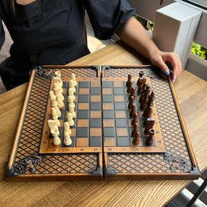 May include: A wooden chess set with a folding board. The board is brown and black with a grid pattern. The chess pieces are light and dark brown. The set is on a wooden table.