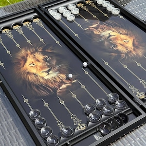 May include: A black backgammon set with a lion's face design. The board features clear and black game pieces, gold accents, and dice. The lion's portrait is in shades of brown and gold.