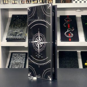 Puede incluir: Caja decorativa negra con un diseño de rosa de los vientos en blanco. La caja está abierta, revelando un interior negro. Otras cajas decorativas son visibles en estantes en el fondo. La caja tiene un acabado brillante.