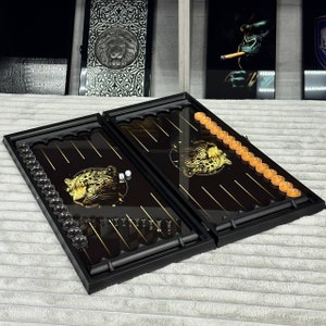 May include: A black backgammon set with a gold leopard design on the playing surface. The set includes clear and orange game pieces, dice, and a black frame. The game is open, ready for play.