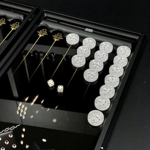 Puede incluir: Un juego de backgammon negro con fichas transparentes con un diseño de reloj. El juego incluye dos dados y elementos decorativos dorados. La superficie de juego es negra brillante.