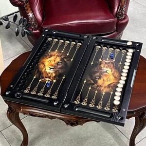 Peut inclure: Un jeu de backgammon noir présentant un portrait de lion détaillé de chaque côté, des repères de points dorés et un ensemble de pions circulaires noirs et blancs. Deux dés bleus sont placés sur la surface du plateau ouvert et plat.