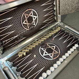 May include: Backgammon board, open, with a black playing surface and silver trim. Each side features a Star of David. Gold and silver game pieces are arranged on the board, ready for play.