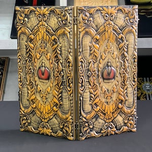 May include: A decorative wooden screen with a gold and black finish. The screen is carved with intricate designs and features a red eye in the center of each panel.
