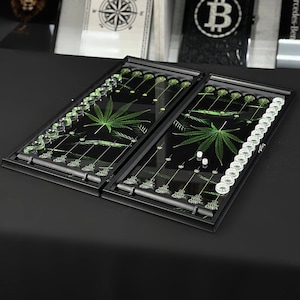 May include: A black backgammon set with a cannabis leaf design. The playing surface is black with green cannabis leaf graphics and clear game pieces. The set includes dice and is displayed on a black surface.