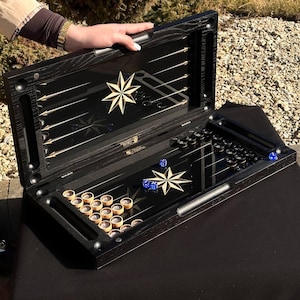 Puede incluir: Un juego de backgammon de madera negra con una superficie de juego de vidrio. El juego está abierto y muestra las fichas y los dados. El juego tiene un diseño de estrella dorada en la superficie de vidrio.