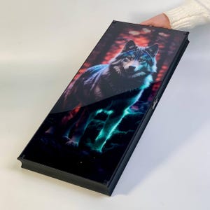 May include: A rectangular black box with a glossy surface featuring a vibrant image of a wolf. The wolf is depicted in a forest setting with red and orange hues in the background and glowing teal accents. The box is held by a person.