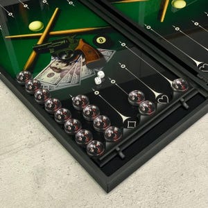 May include: A black and green backgammon board with a pool table design. The board features a revolver, money, dice, and pool balls. The backgammon pieces are clear with red centers. The board is open and ready to play.