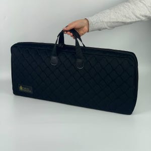 May include: Black rectangular case with a quilted pattern and a handle. The case has a zipper closure and a small gold label with text. The case is held by a person.
