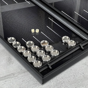 May include: A black backgammon set with a textured surface, featuring silver-coloured game pieces and two dice. The set is open, displaying the playing field and the arrangement of the pieces.