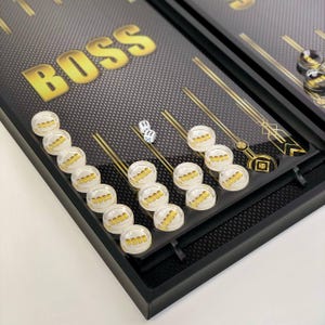 Puede incluir: Un juego de backgammon negro y dorado con la palabra "BOSS" impresa en el tablero. Las fichas del juego son blancas con letras doradas. Dos dados blancos están en el tablero.