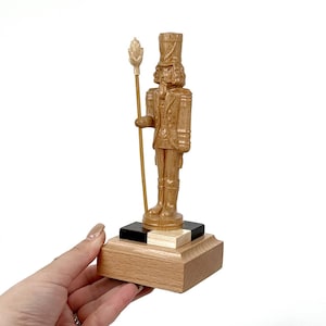 Pode incluir: Figura de quebra-nozes de madeira com uniforme de soldado, segurando um bastão com um topo decorativo. A figura castanho-claro tem um design detalhado, em uma base de madeira com detalhes em preto e branco.