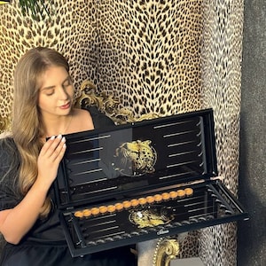 May include: A black backgammon set with gold accents, featuring a decorative leopard design. The set is open, revealing the playing surface and game pieces. The background has a leopard print.