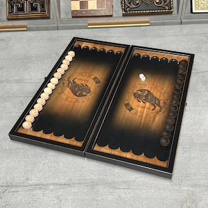 May include: A wooden backgammon board with a black frame and a dark brown playing surface. The board features the Taurus zodiac symbol and comes with light-colored game pieces and dice. The board is open, ready for play.