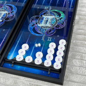 Puede incluir: Tablero de backgammon con un diseño celestial. El tablero presenta un fondo azul con un patrón de estrellas y un símbolo del zodiaco Géminis. Fichas blancas con un símbolo de Géminis están dispuestas en el tablero, junto con dos dados. El tablero está enmarcado en negro.