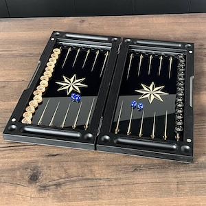 May include: A black backgammon board featuring a glossy interior with gold-colored triangular points and a central star design. The set includes light-colored and dark-colored circular checkers arranged along the sides and four blue dice.