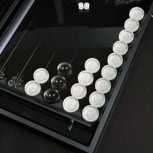 May include: A black glass game board with white and black marbles. The board has a series of lines and a decorative design. Two white dice are visible in the upper right corner of the image.