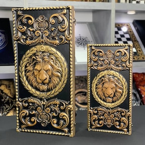 Puede incluir: Dos cajas de madera con adornos dorados ornamentados y tallas de cabeza de león. La caja más grande es negra con una cabeza de león dorada en una corona, mientras que la caja más pequeña es marrón con una cabeza de león dorada en una corona.