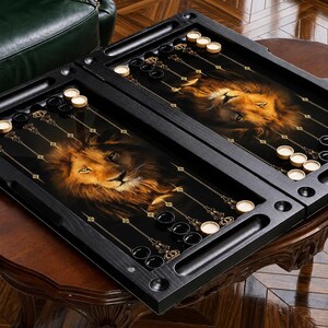 Peut inclure: Un plateau de backgammon en bois noir présentant un graphique détaillé de visage de lion sur la surface de jeu. Le plateau comprend des marqueurs de points dorés et un ensemble de pions circulaires noirs et blancs disposés sur l'intérieur.