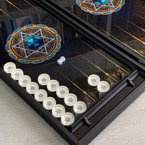 May include: A black backgammon game board with gold accents and a blue and gold star design. The board is open and shows white playing pieces.