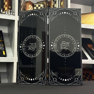May include: Two black decorative panels with white celestial and zodiac designs. Each panel features a central circular zodiac symbol, surrounded by astrological symbols and phases of the moon. The panels are framed with a white border.