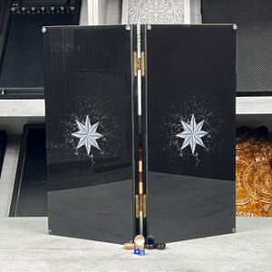 May include: A black backgammon board with a starburst design. The board is open, revealing the playing surface and game pieces. The board is made of a glossy material and has gold hinges.