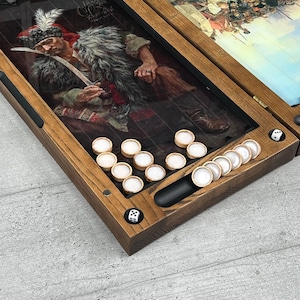 May include: Wooden backgammon set with a detailed painting of a historical figure. The set includes white and tan game pieces, dice, and a black storage area. The wooden frame has a natural finish.