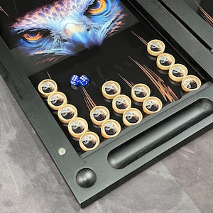 May include: A black backgammon board featuring an artistic rendering of an owl's face in shades of blue and orange. The board includes wooden game pieces with owl imagery and two blue dice. The board is framed in black.
